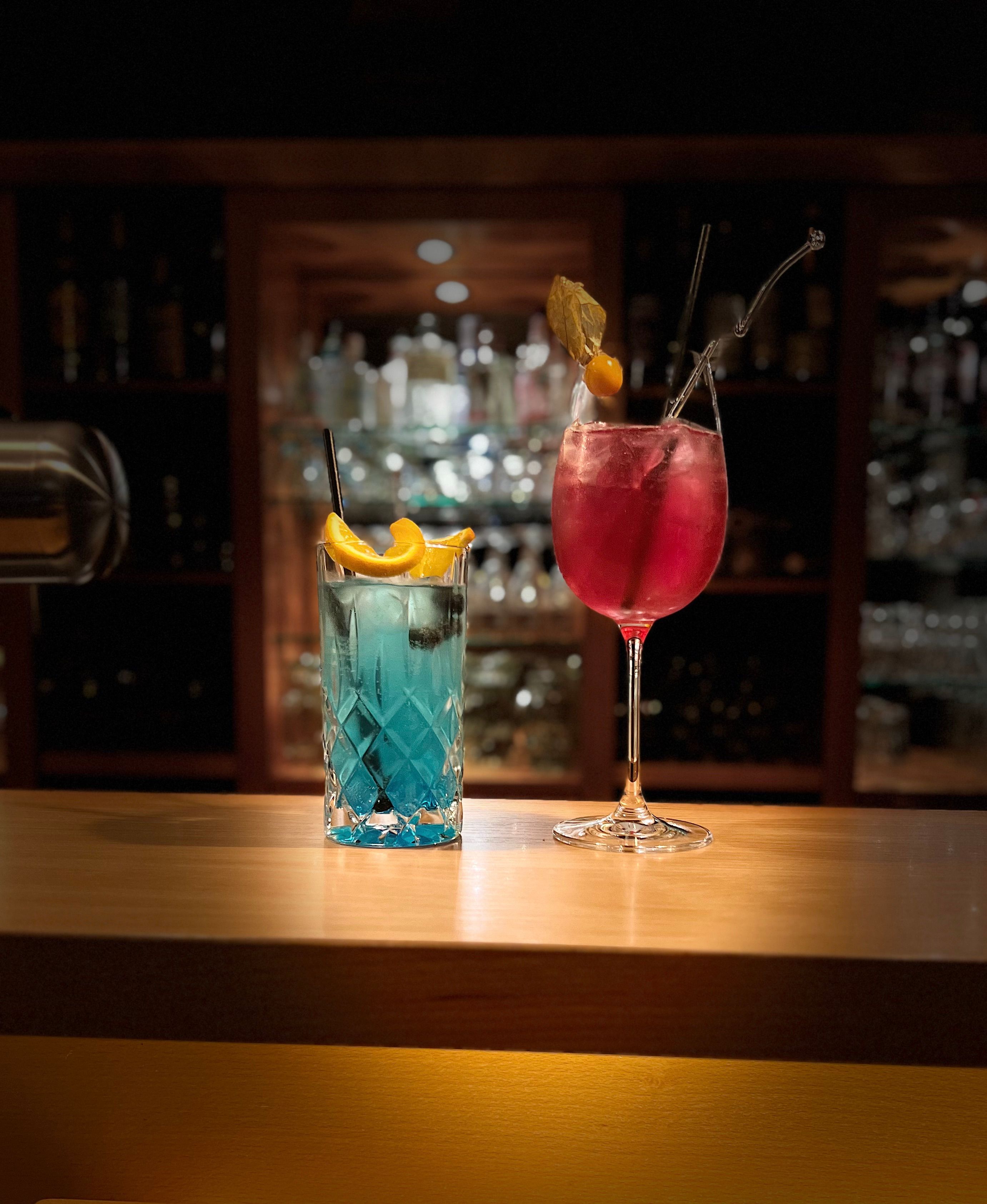 Zwei Cocktails auf der Bartheke — Blue Drink und Pink Spritz vor der beleuchteten Flaschenwand